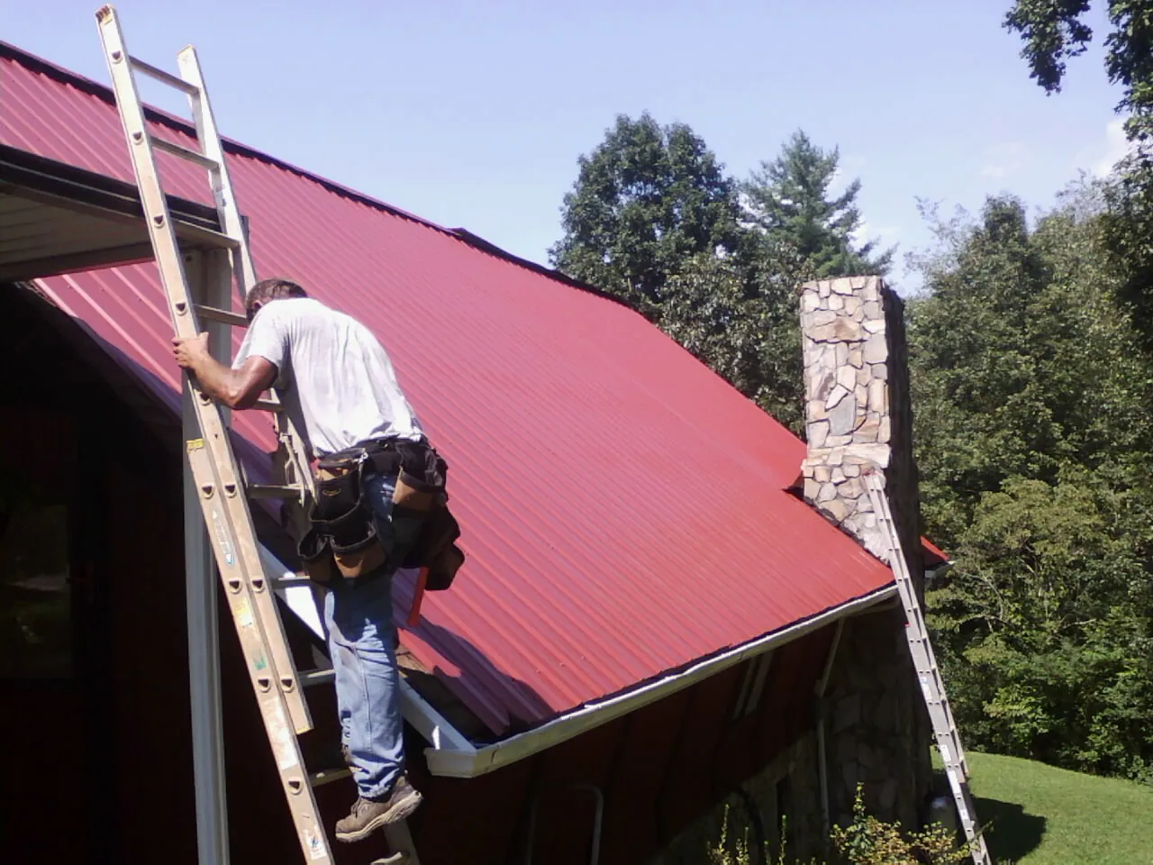 Roofer on a ladder beside a red metal roof and stone chimney — Soffit & Fascia Repair by Roofing Brothers in Nodaway